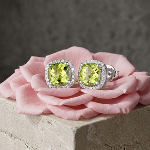 A pair of cushion-cut  peridot green stud earrings with diamond halo settings in silver, displayed on a pink rose against a textured grey background.