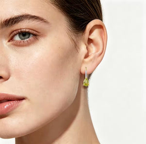 A close-up of a model's ear wearing a delicate gold dangle earring with a pear-cut green peridot and diamond gemstone and a small iridescent accent.