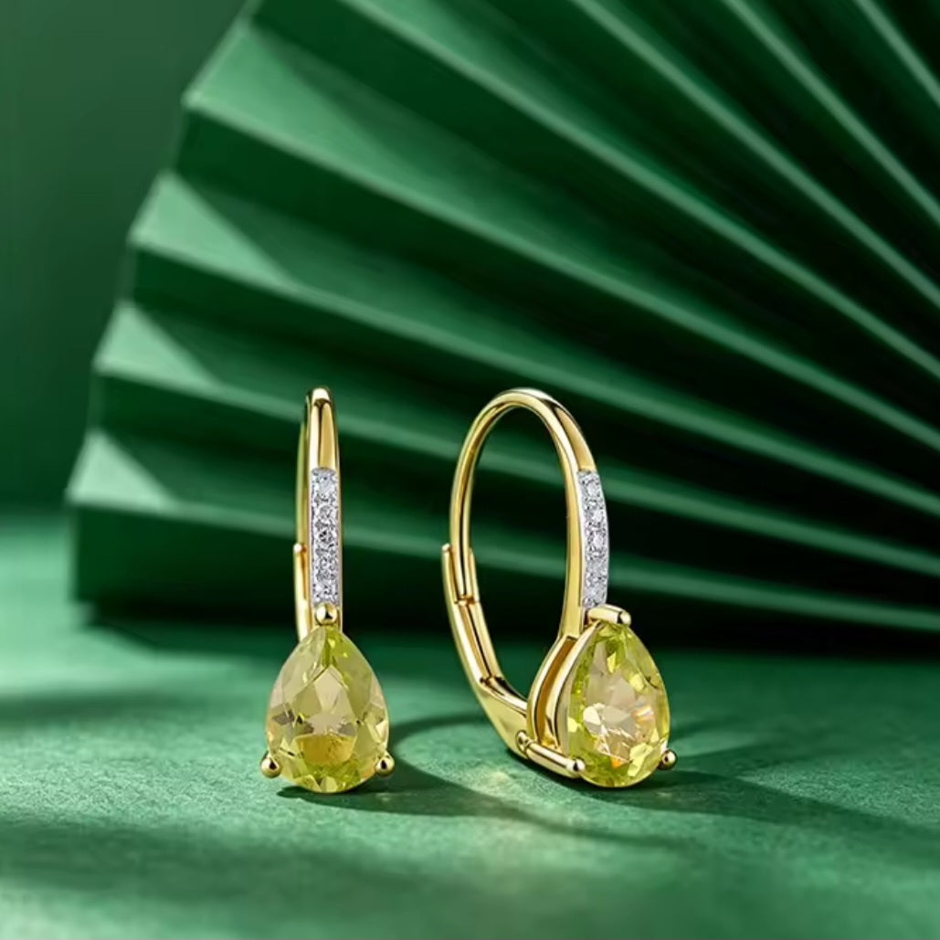 A pair of gold hoop earrings with pear-cut peridot gemstones and small clear diamond stones along the hoops, displayed on a green surface with a matching green fan-shaped backdrop.