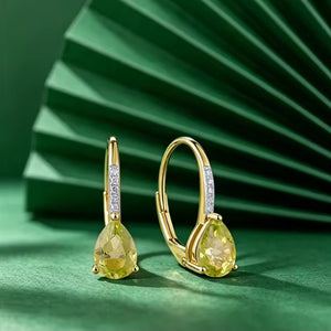 A pair of gold hoop earrings with pear-cut peridot gemstones and small clear diamond stones along the hoops, displayed on a green surface with a matching green fan-shaped backdrop.