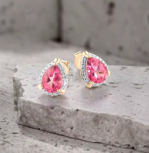 A pair of gold teardrop-shaped stud earrings with large pink natural topaz gemstones and small clear accent natural diamond stones, displayed on a textured grey stone surface.