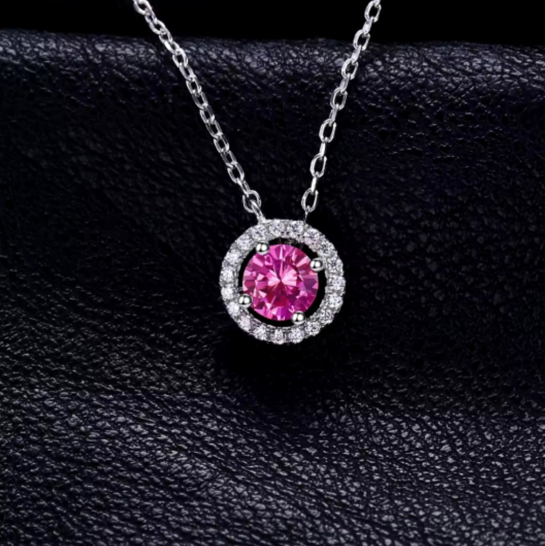 A silver necklace featuring a central hot pink sapphire gemstone surrounded by smaller clear sapphires, displayed against a black background.