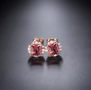 A pair of rose gold stud earrings featuring cushion-cut pink gemstones surrounded by small clear accent stones, displayed on a dark gray surface.