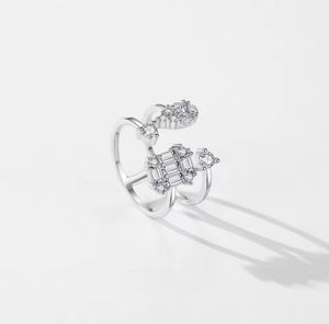 A silver open ring ring featuring a central cluster of baguette and round-cut clear gemstones, with smaller floral clusters at the top and side, photographed on a clean white background with soft shadows.