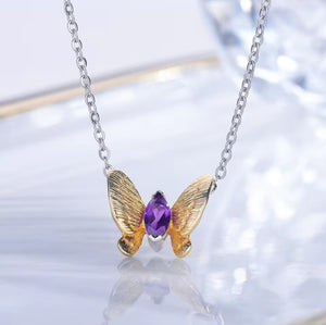 A butterfly-shaped pendant necklace with textured gold wings and a central purple gemstone, suspended from a silver link chain on a reflective white surface.