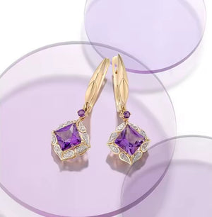A pair of gold lever back earrings with large purple amethyst square-cut gemstones, accented by small round purple amethyst stones and natural diamonds, displayed on a purple circular stand against a white background.