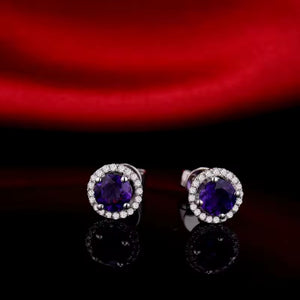 A pair of oval stud earrings with deep purple natural amethyst center stones surrounded by clear halo diamond stones in a silver-toned setting, displayed on a reflective black surface with a red satin background.