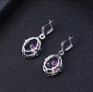 A pair of silver drop earrings with faceted iridescent natural mystic quartz -purple and green stones, displayed on a textured black surface.