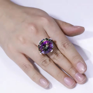 A close-up of a hand wearing a large, round natural mystic quartz gemstone ring with a purple and green colour-shifting effect on a white background.
