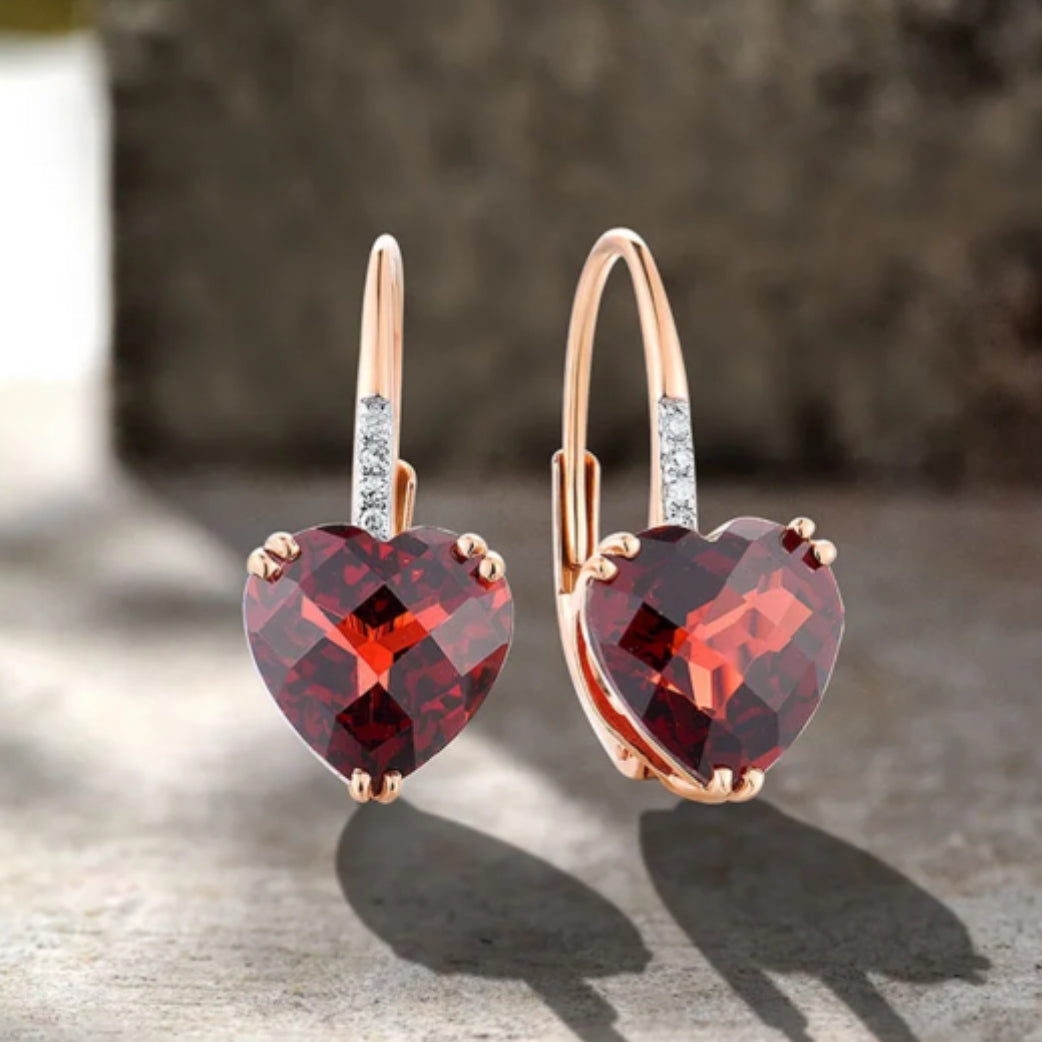 A pair of rose gold heart-shaped earrings with one larger garnet stone and smaller diamonds, set in rose gold bands.