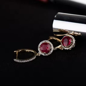 A pair of gold hoop earrings with oval red natural ruby gemstones surrounded by small clear stones, displayed on a black surface with a silver cylindrical prop.