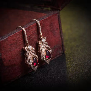A pair of rose gold drop earrings with clear pavé stones and a deep red teardrop gemstone, displayed on a dark reddish-brown wooden box.