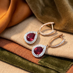 A pair of gold hoop earrings with teardrop-shaped red ruby gemstones surrounded by small diamond clear stones, displayed on a textured fabric background with warm brown and olive tones.