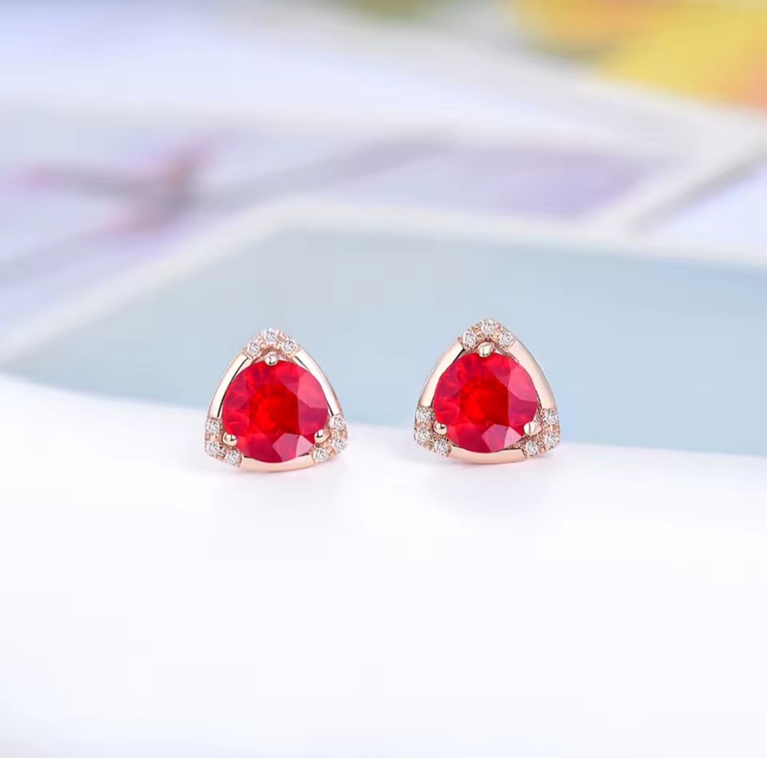 A pair of triangular stud earrings with faceted red ruby gemstones and small clear diamond stones set in rose gold, displayed on a clean white surface with a soft-focus background.