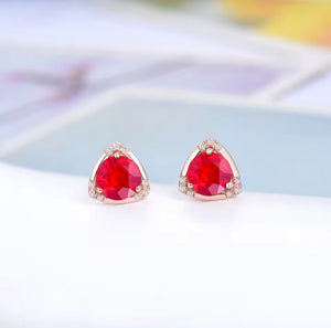 A pair of triangular stud earrings with faceted red ruby gemstones and small clear diamond stones set in rose gold, displayed on a clean white surface with a soft-focus background.