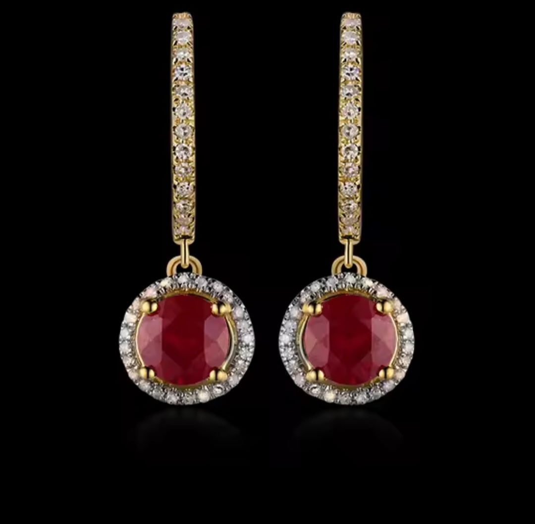 A pair of gold huggie drop earrings featuring round-cut deep red ruby centers surrounded by a halo of small clear stones, with the huggie bands paved with clear stones, set against a solid black background.
