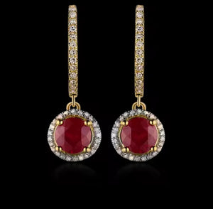 A pair of gold huggie drop earrings featuring round-cut deep red ruby centers surrounded by a halo of small clear stones, with the huggie bands paved with clear stones, set against a solid black background.