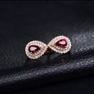 A pair of teardrop-shaped earrings with deep red ruby gemstones surrounded by small clear diamond stones in a rose gold setting, displayed on a textured black surface.