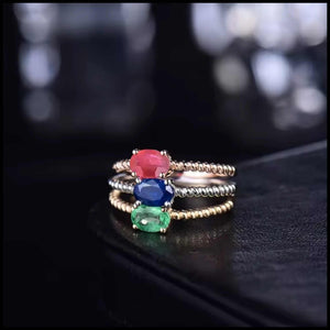 Stacked 14k gold rings with oval ruby, sapphire, and emerald gemstones on black surface