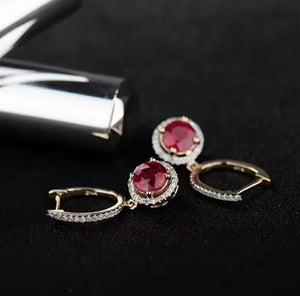 A pair of gold hoop earrings with oval red ruby gemstones surrounded by small clear stones, displayed on a black textured surface next to a silver cylindrical object.