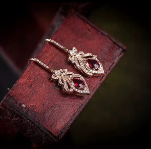 A pair of rose gold dangle earrings with red teardrop-shaped gemstones and small clear stones, displayed on a dark reddish-brown wooden block against a dark blurred background.