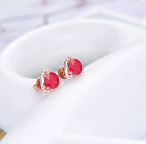 A pair of rose gold stud earrings with oval-cut red ruby gemstones and small clear diamond accent stones, displayed on a white ceramic stand.