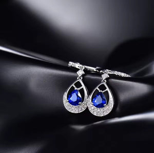 A pair of white gold teardrop dangle earrings with deep blue sapphire teardrop gemstones and small clear stones, displayed on a black satin fabric background.