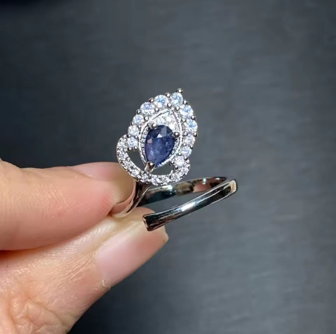 A close-up of a hand holding a silver-toned ring with a central oval blue sapphire surrounded by a halo of small clear stones, set against a dark gray background.