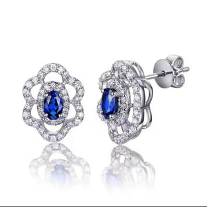 A pair of white gold stud earrings with oval-cut blue centre sapphire stones and clear natural diamond accent stones, shown on a white background with a soft reflection below.