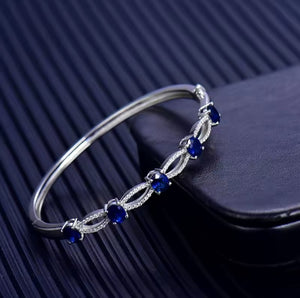 A white gold bangle bracelet with five round-cut deep blue sapphire gemstones, accented by small clear diamonds, resting on a dark textured surface next to a black leather-like block.