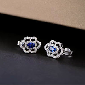 A pair of  white gold shaped stud earrings with oval blue centre stones and clear surrounding natural diamond stones, displayed on a dark surface with a beige textured background.