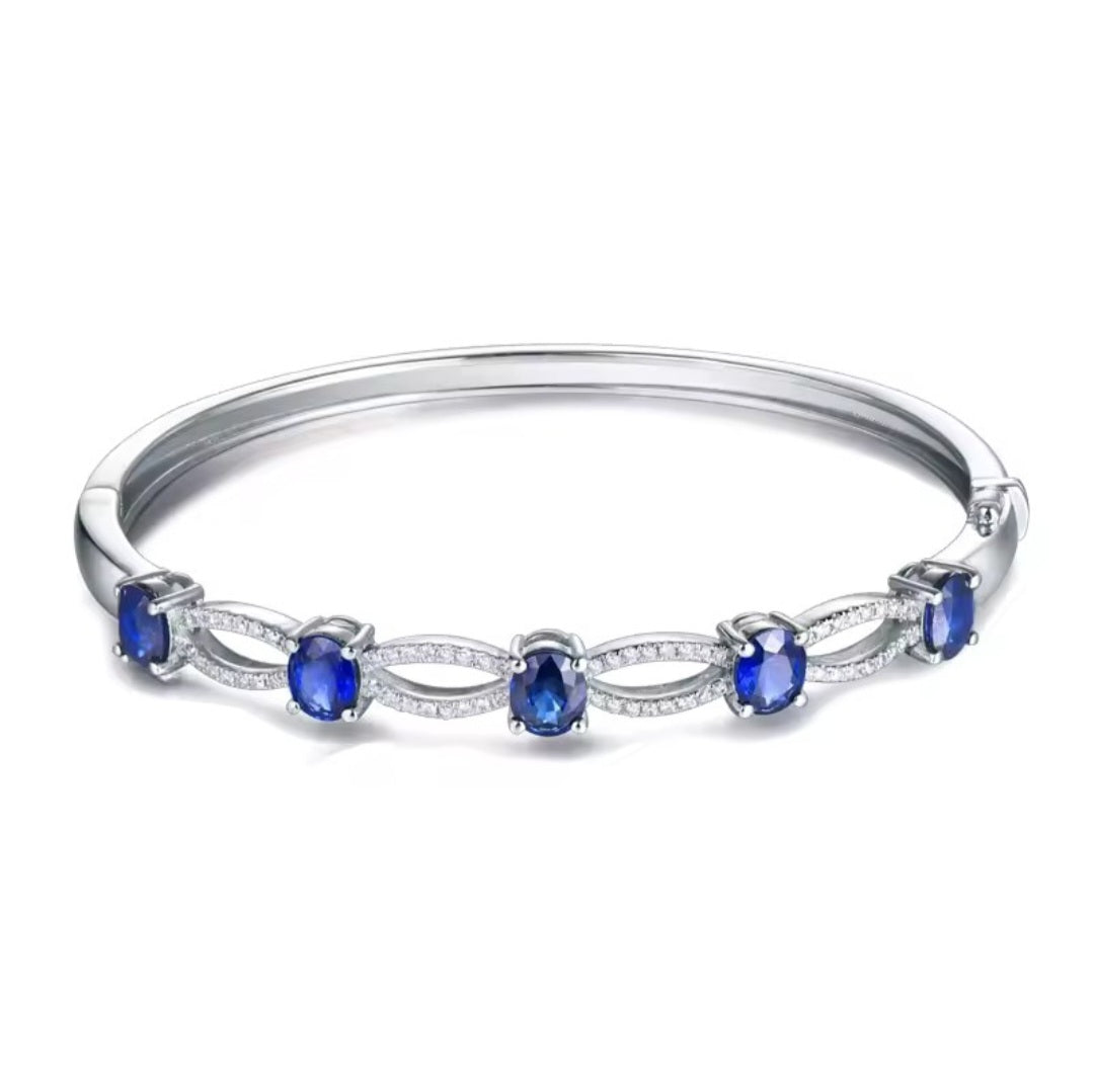 A white gold bangle bracelet featuring five oval-cut deep blue sapphires and small clear pavé stones, set against a plain white background.