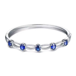 A white gold bangle bracelet featuring five oval-cut deep blue sapphires and small clear pavé stones, set against a plain white background.