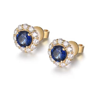 A pair of gold post earrings featuring a round deep blue sapphire center stone surrounded by a halo of small clear round-cut diamond stones, shown on a clean white background.