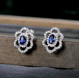 A pair of white gold flower-shaped stud earrings with oval blue gemstones and clear accent natural diamond stones, displayed on a dark textured surface.