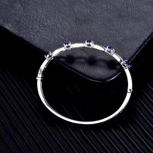 A  white gold circular bangle bracelet with small blue sapphire gemstone accents, displayed on a textured black surface.