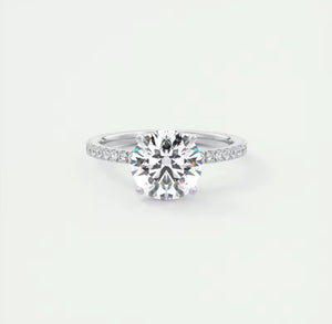 A classic solitaire engagement ring featuring a round brilliant-cut center stone set on a polished silver-colored band with pavé-set accent stones, shown against a clean, light off-white background.