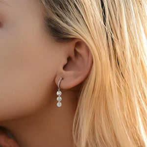 A close-up of a model's ear wearing a small silver huggie hoop earring with three dangling clear gemstones.