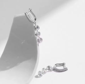 A pair of silver-toned huggie hoop earrings with a dangling cluster of clear round-cut gemstones, displayed on a white jewelry stand against a bright white background.