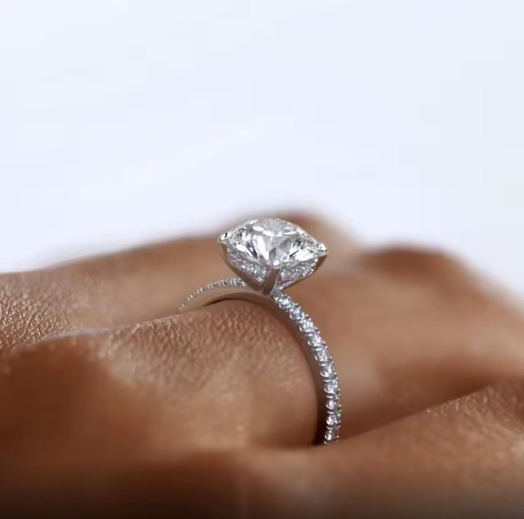 A close-up of a silver moissanite ring with a round-cut centre stone and a pave-set band, worn on a finger against a soft light background.