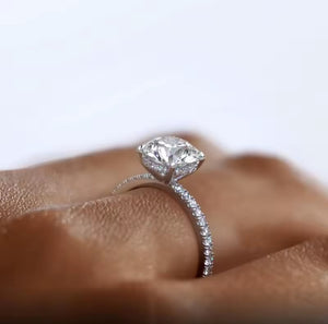 A close-up of a silver moissanite ring with a round-cut centre stone and a pave-set band, worn on a finger against a soft light background.