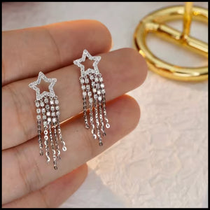 Diamond star-shaped earrings with dangling chains held between fingers against a light background.