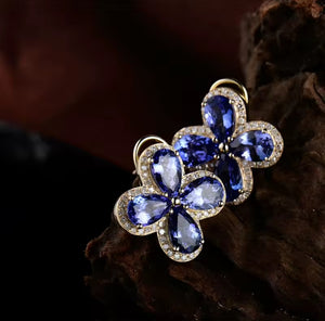 A pair of gold flower-shaped rings with deep blue teardrop tanzanite gemstones and small clear natural diamond accent stones, displayed on a dark textured wooden surface.