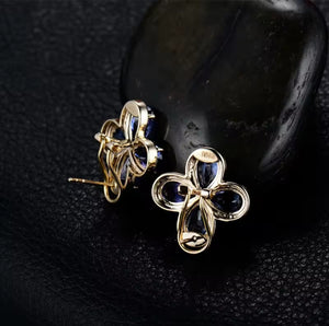 A pair of gold four-petal flower stud earrings with dark blue inlay, displayed on a black textured surface next to a polished black stone.