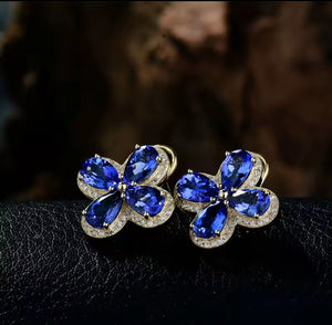 A pair of yellow gold flower-shaped , clover earrings featuring large blue tanzanite gemstones and small clear diamond stones in a halo setting, displayed on a dark textured surface with a blurred dark brown background.
