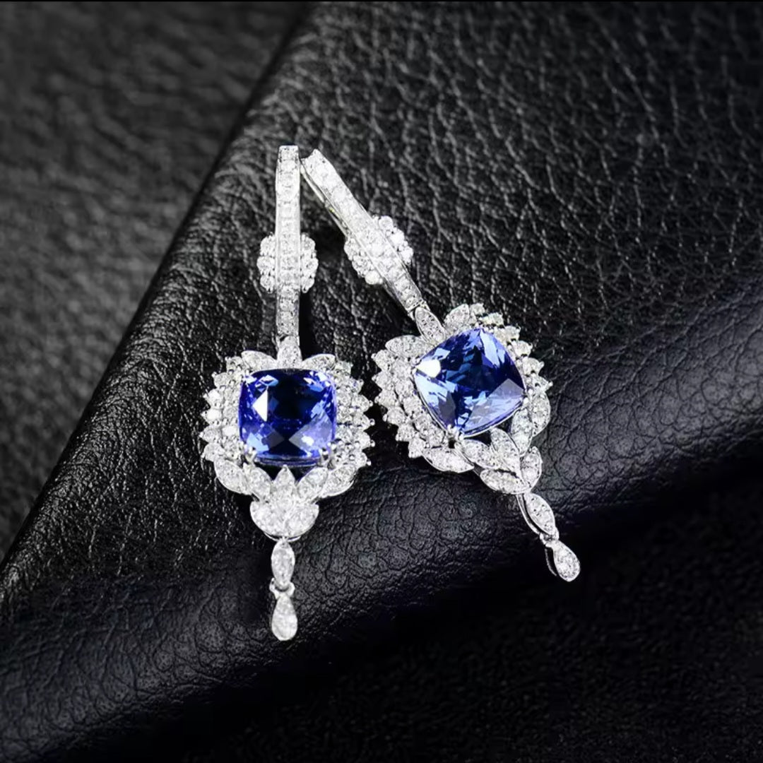 A pair of white gold drop earrings featuring large square-cut blue tanzanite gemstones surrounded by small clear diamond stones, displayed on a textured black leather surface.