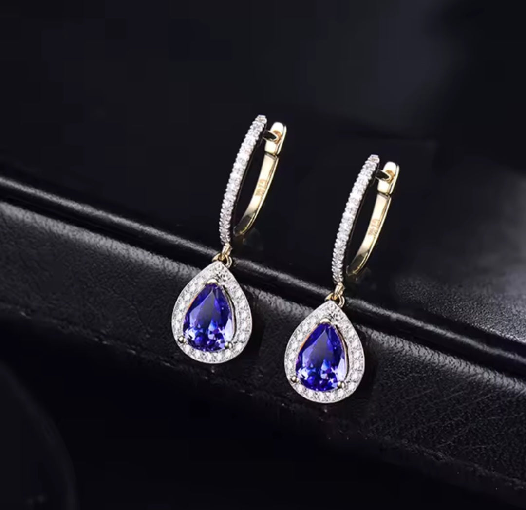 A pair of gold huggie hoop earrings with teardrop-shaped blue tanzanite gemstone pendants, accented with clear pavé diamond stones, displayed on a black leather surface.