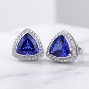 A pair of triangular stud earrings featuring deep blue marquise-cut center stones surrounded by silver-toned halos of small clear stones, displayed on a light marble surface.