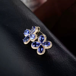 A pair of gold flower-shaped stud earrings with deep blue tanzanite gemstones and clear  diamond accent stones, resting on a black textured surface.
