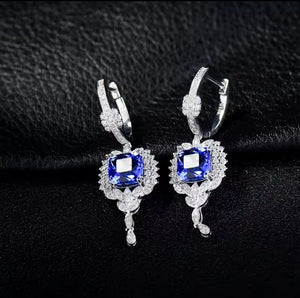 A pair of white gold earrings with rectangular blue tanzanite gemstones and surrounding clear crystal detailing, displayed on a textured black background.